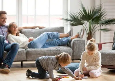 Lebensversicherung: Junge Eltern entspannen zu Hause auf dem Sofa, während die beiden Kinder auf dem Boden spielen