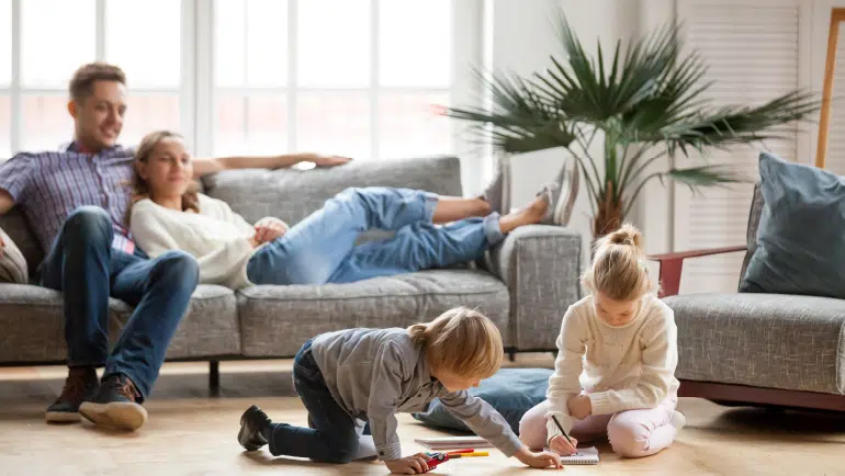Lebensversicherung: Junge Eltern entspannen zu Hause auf dem Sofa, während die beiden Kinder auf dem Boden spielen