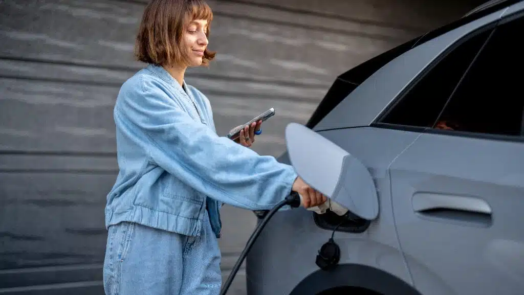 Bild einer Frau, die gerade den Stecker in ihren Elektro Kleinwagen zum Laden steckt, während Sie ihr Handy in der Hand hält.
