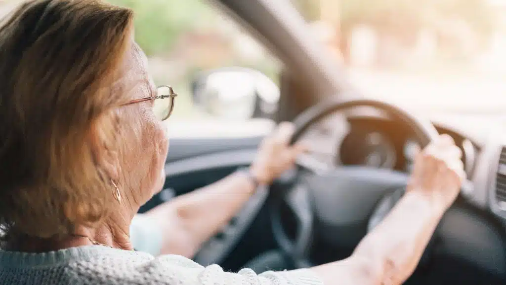 Foto einer älteren Frau beim Autofahren, seitlich von den mittleren Sitzen aus fotografiert.