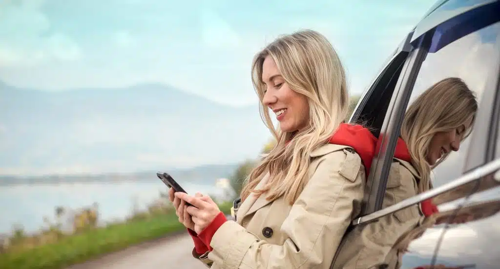 Eine junge Frau steht angelehnt an ein Auto und bedient ein Smartphone