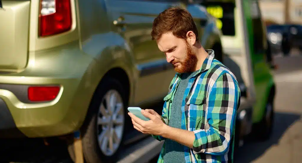Junger Mann mit Smartphone in der Hand steht vor einem Auto auf einem Abschleppwagen