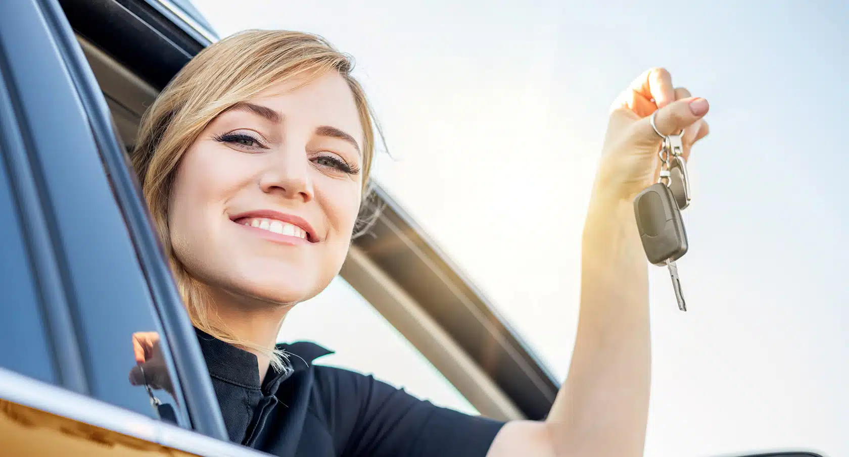 Lächelnde junge Frau schaut aus einem Autofenster und zeigt einen Autoschlüssel