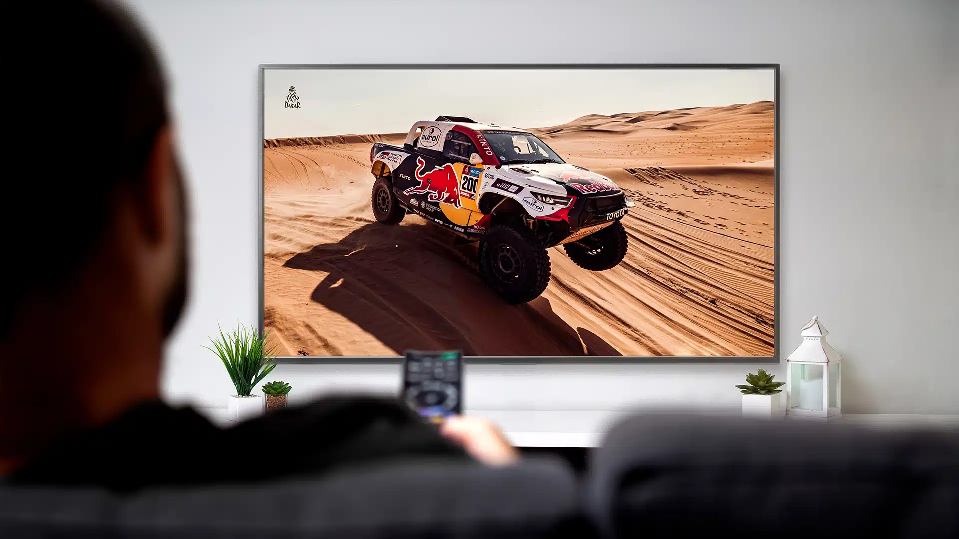 Blog-Titelbild-Die besten Autoserien (1) Autoserien im Überblick: Ein Mann mit Fernbedienung in der Hand, der sich eine Auto Wüsten-Rallye im TV ansieht.