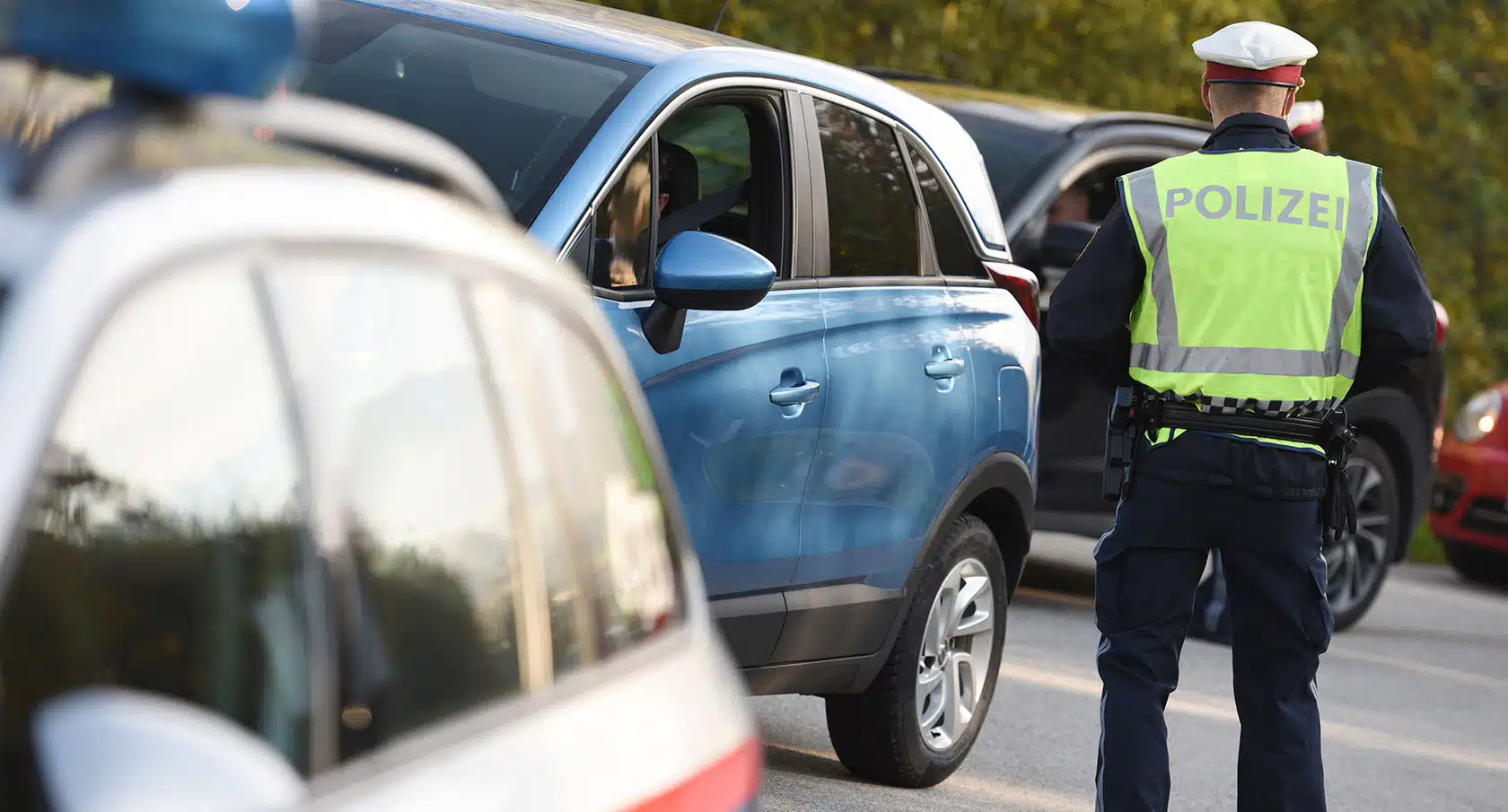 Foto von einer Grenzkontrolle, bei der ein Polizist von hinten zu sehen ist, der neben einem blauen Auto steht.