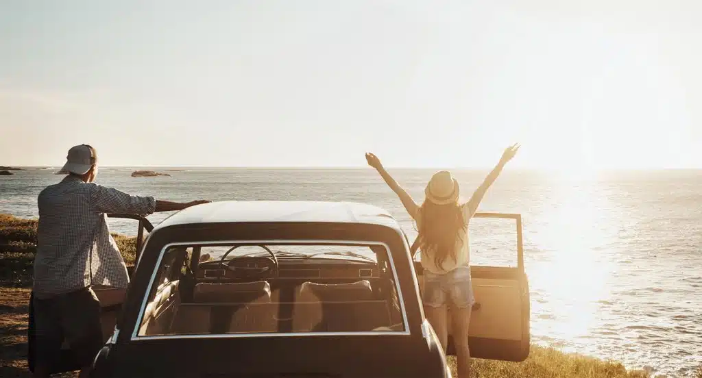 Ein Auto steht am Strand, daneben ein junger Mann und eine junge Frau