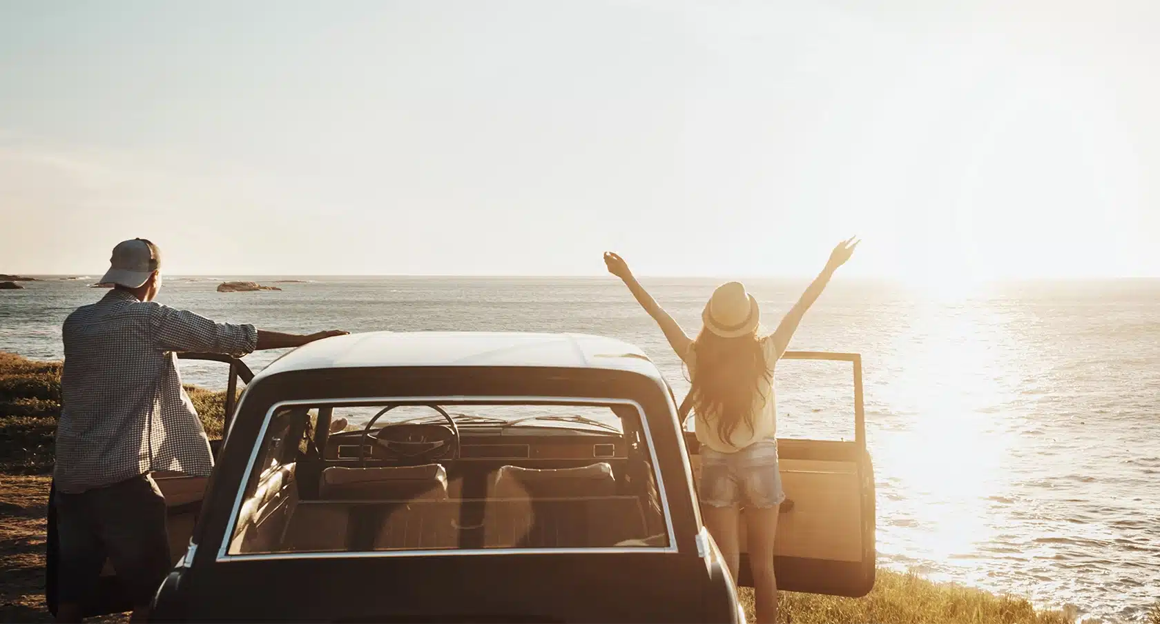 Ein Auto steht am Strand, daneben ein junger Mann und eine junge Frau