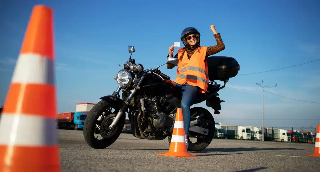 Motorradfahrer in Warnweste sitzt auf einem Motorrad und winkt. Im Vordergrund stehen Verkehrshütchen.