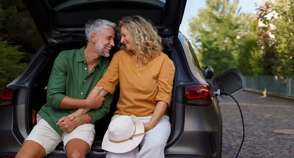 Pärchen sitzt auf der Ladekante eines geöffneten Kofferraums von einem E-Auto beim Ladevorgang und hält Händchen
