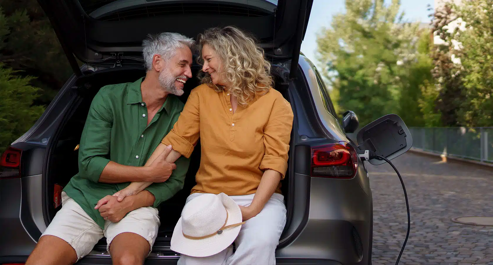 Pärchen sitzt auf der Ladekante eines geöffneten Kofferraums von einem E-Auto beim Ladevorgang und hält Händchen