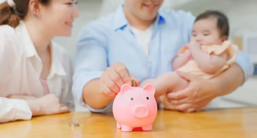 Vater und Mutter sitzen mit Baby im Arm an einem Tisch und stecken eine Münze in ein Sparschwein