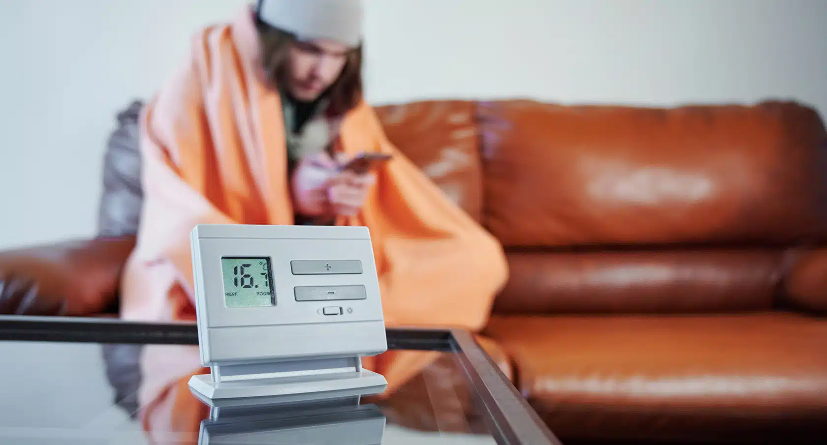 Junger Mann sitzt frierend und in eine Decke gewickelt auf einem Sofa vor einem Thermometer