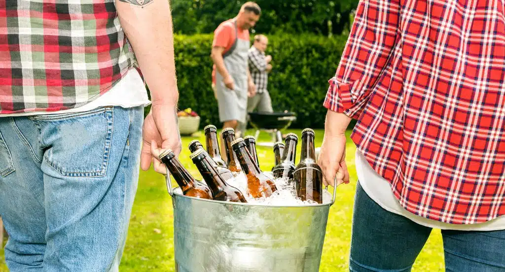 Zwei Männer tragen einen Kübel mit Eiswürfeln und Bierflaschen zu einem Grill im Garten