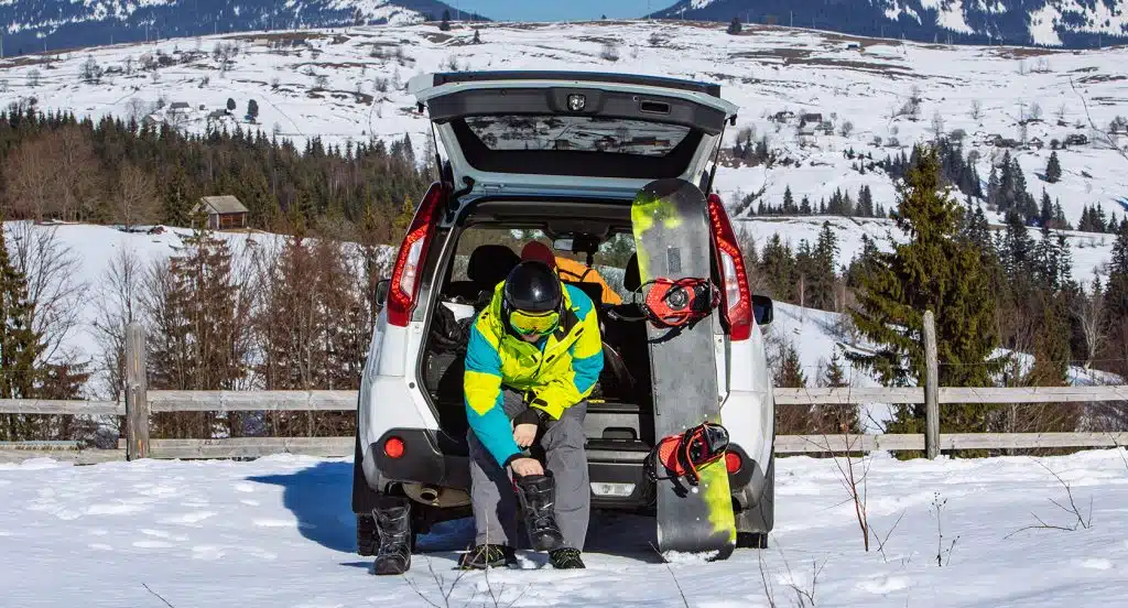 Snowboardfahrer sitzt auf der Laderampe eines geöffneten Kofferraums und wechselt die Schuhe