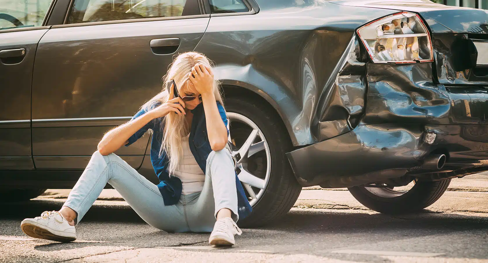 Junge Frau sitzt vor einem zerbeulten Auto und telefoniert
