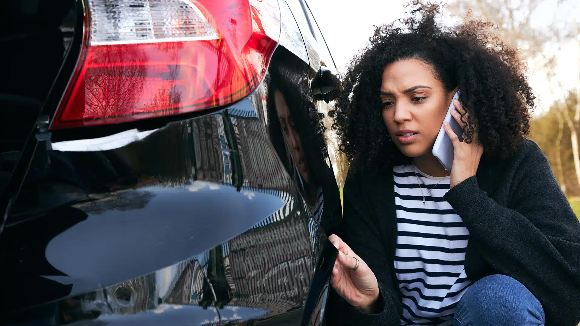 Eine Frau kniet neben einem angefahrenen Auto mit Schramme und telefoniert mit dem Smartphone