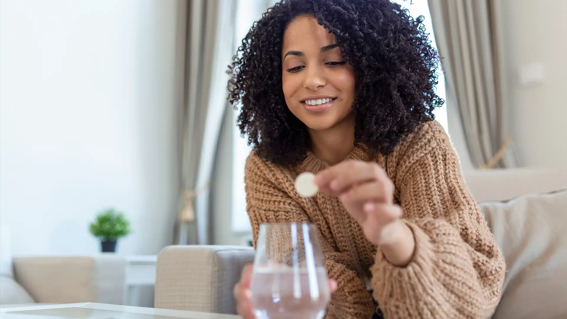 Lächelnde Frau hält eine Brausetablette zwischen den Fingern, vor ihr steht ein Wasserglas.