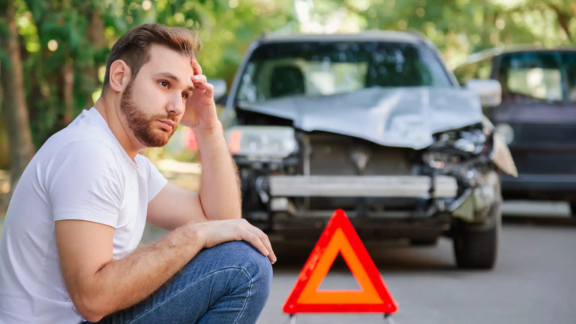 Mann sitzt nach einem Unfall auf dem Bürgersteig und hat gestresst die Hand an den Kopf gelegt. Hinter ihm ein demoliertes Auto und ein Warndreieck.