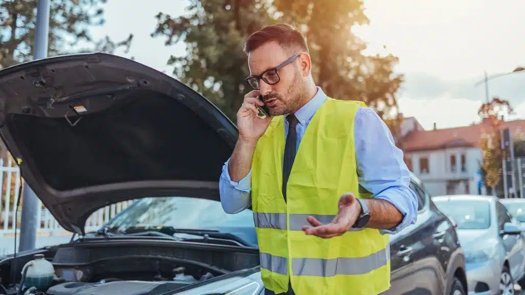 Ein Mann in einer Warnweste steht telefonierend vor der geöffneten Motorhaube eines Autos