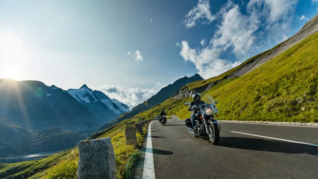 Motorradfahrer fährt eine Straße an einem Hügel entlang. Im Hintergrund sind Berge.