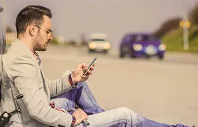 Mann sitz auf dem Boden an sein Auto gelehnt und tippt auf sein Handy