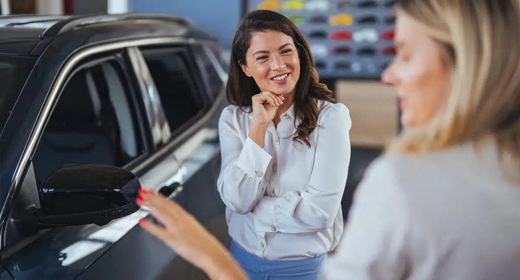 Eine Frau erklärt einer anderen Frau in einer Werkstatt vor einem Auto etwas