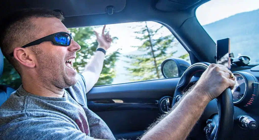 Junger Mann mit Sonnenbrille hinterm Steuer eines Autos hält eine Hand aus dem Fenster des Autos und lacht