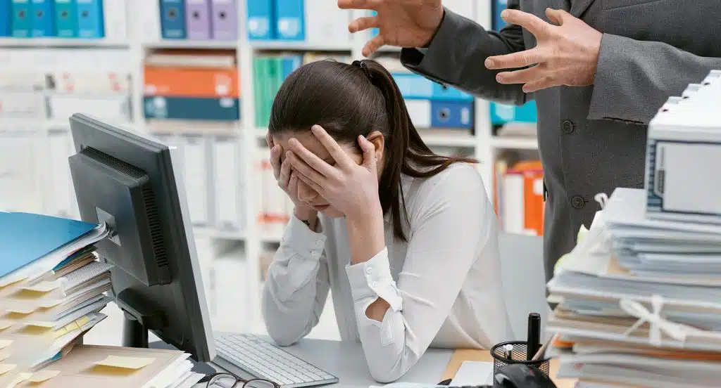 Eine frustrierte Frau schlägt vor einem Computer die Hände vor ihr Gesicht, während hinter ihr zwei Hände hysterisch gestikulieren