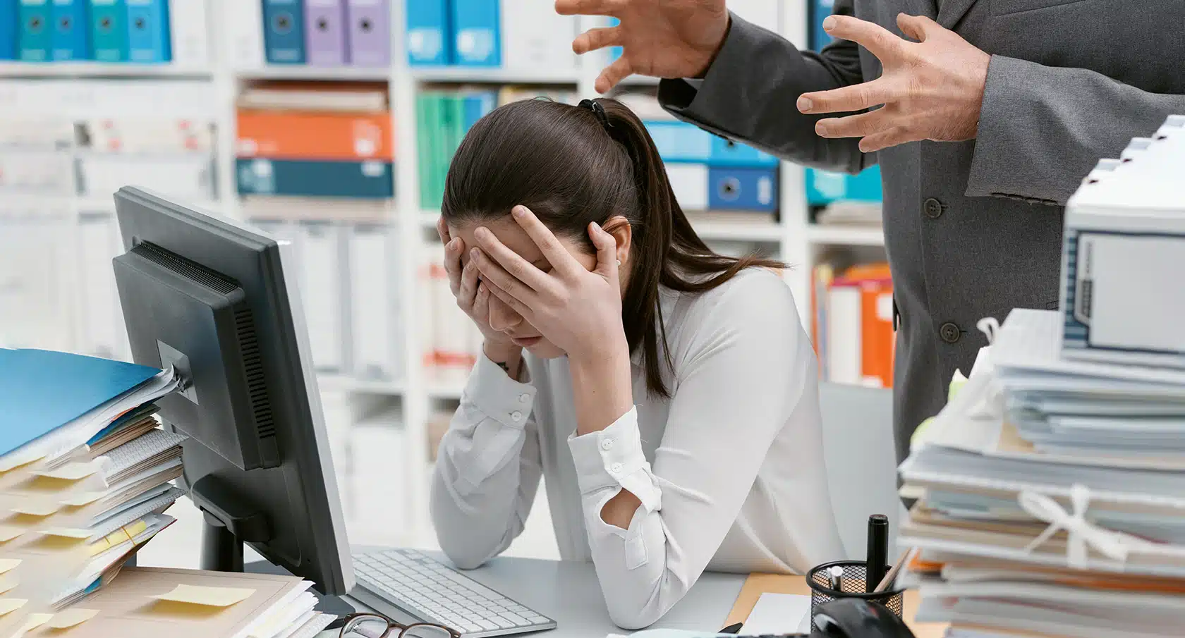 Eine frustrierte Frau schlägt vor einem Computer die Hände vor ihr Gesicht, während hinter ihr zwei Hände hysterisch gestikulieren