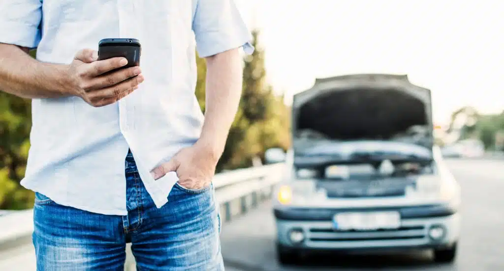 Autofahrer mit Smartphone steht vor seinem Fahrzeug und ruft Hilfe